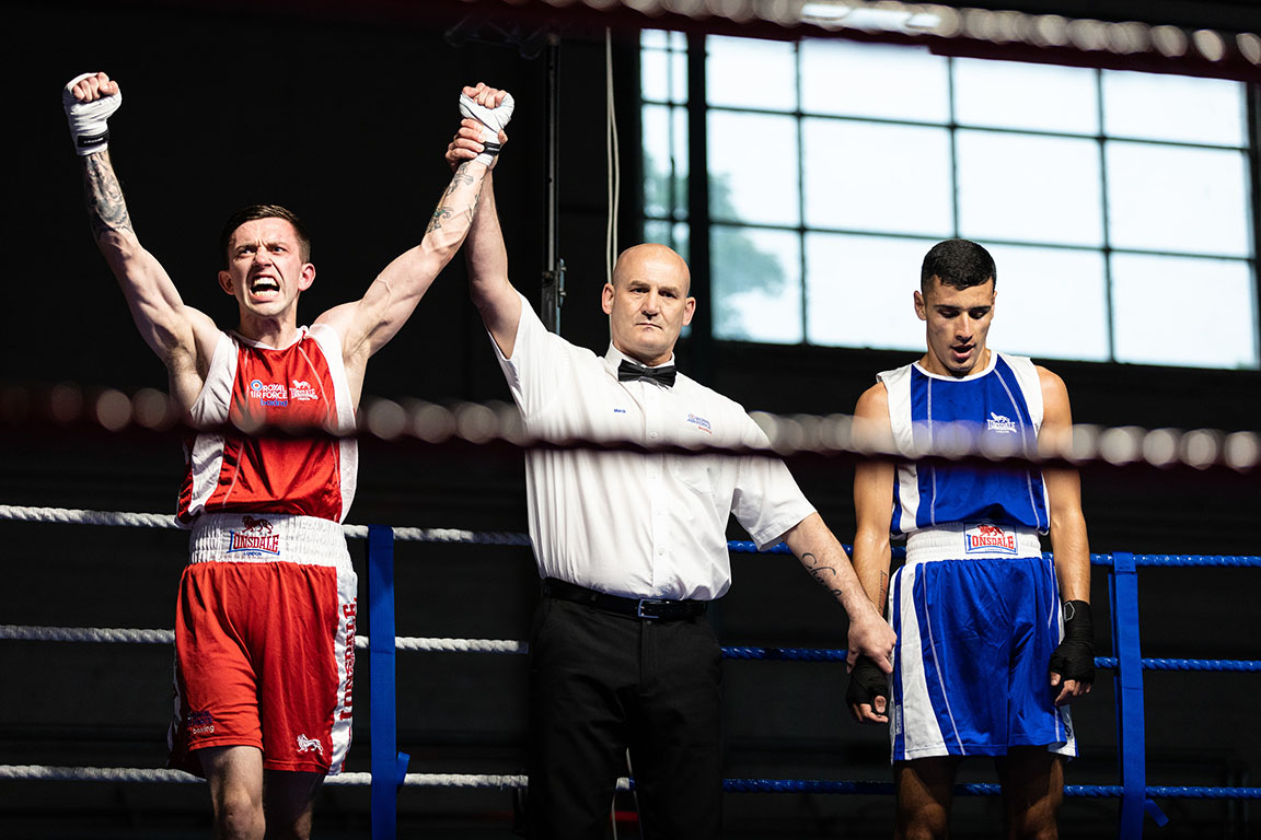 Image shows RAF aviators in a boxing ring, as the referee holding up the arm of the winner in red.